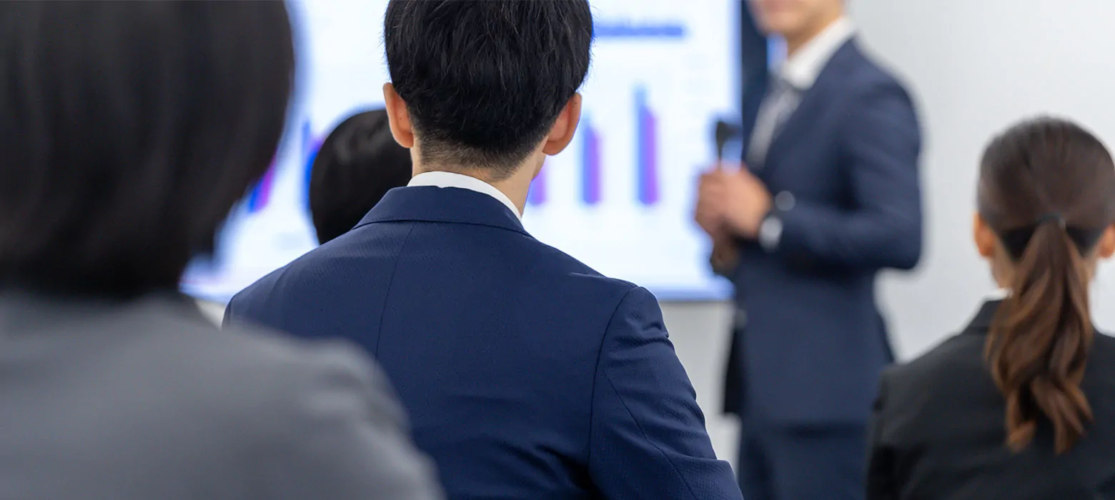 スーツ姿の参加者が背後から講師のプレゼンテーションを聴講するセミナー会場の様子