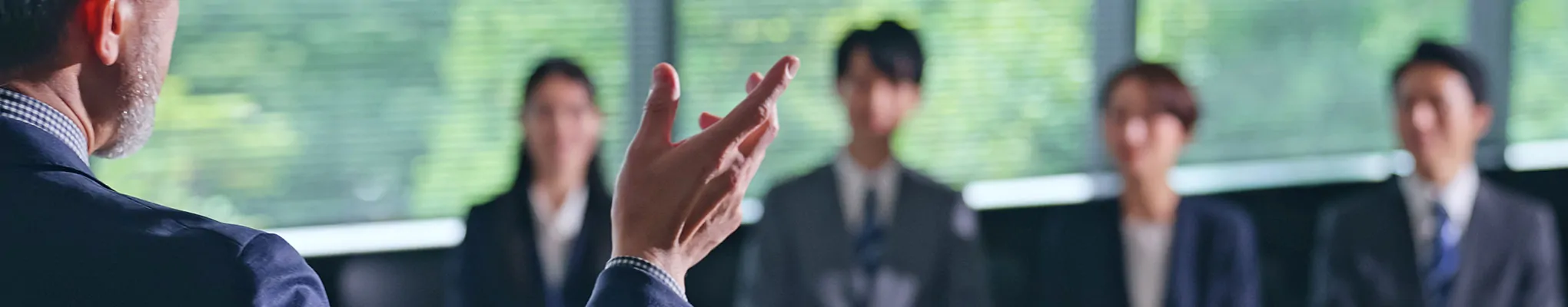 セミナー会場で、背後から手を挙げて説明する講師と、スーツ姿の参加者が座って話を聞く様子