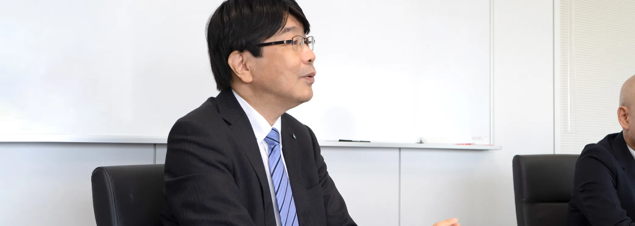 白い会議室でスーツ姿の男性が横顔で話すインタビューの様子
