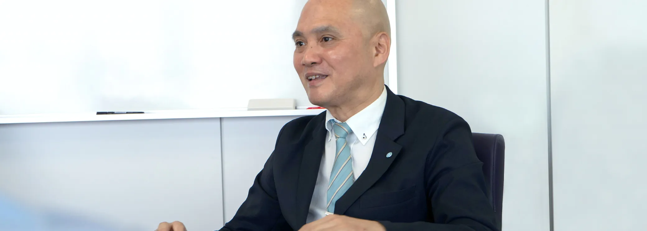 白い会議室でスーツ姿の男性がテーブル越しに笑顔で話すインタビューの様子