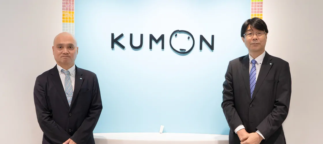 KUMONのロゴを背景に、ダークスーツを着た二名の男性がカウンター両脇でポーズをとっている