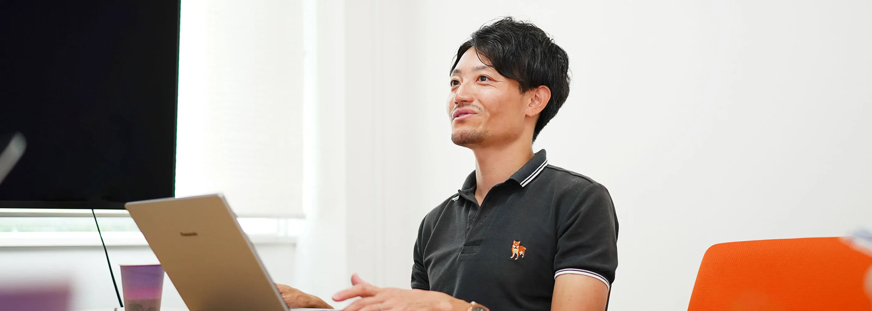 会議テーブルで手がノートPCを操作し、手前にぼけたコーヒーカップが映るインタビューシーン
