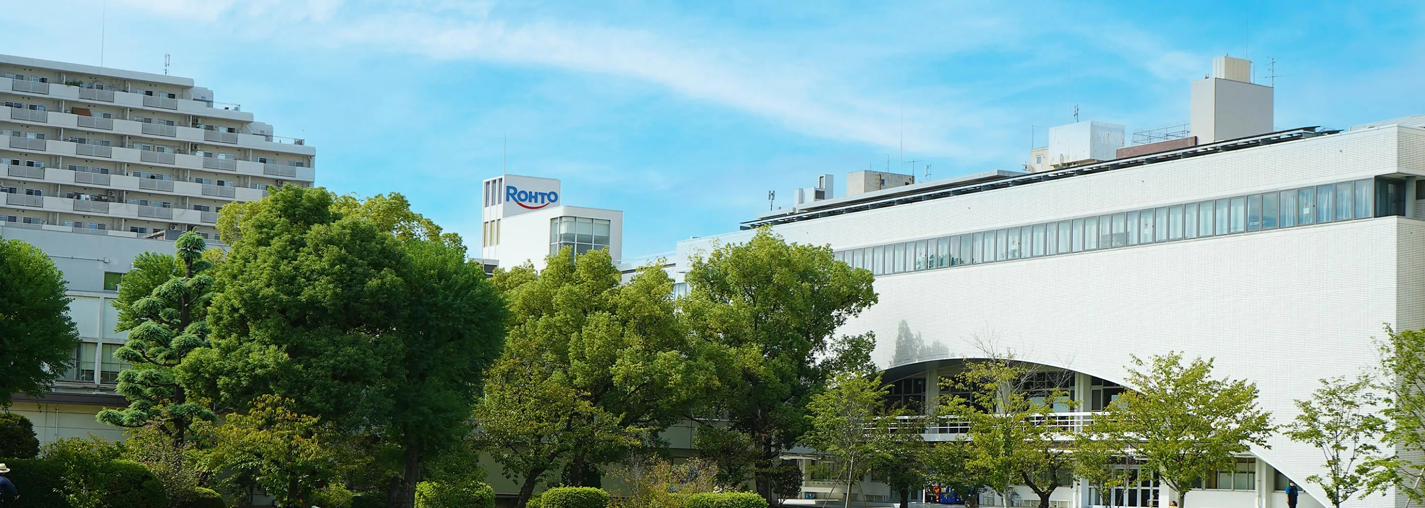 緑の木々の向こうにROHTOロゴが掲げられた白い社屋と青空の風景。