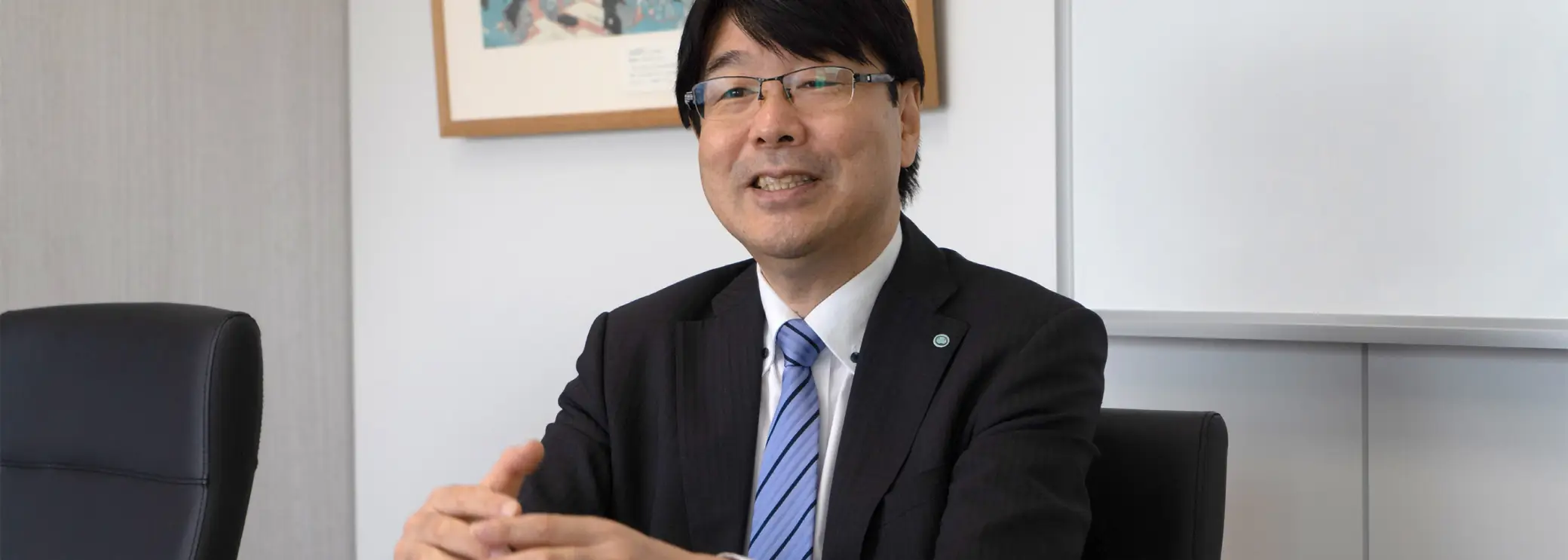 白い会議室でスーツ姿の男性が微笑みながら身ぶりを交えて話すインタビュー風景