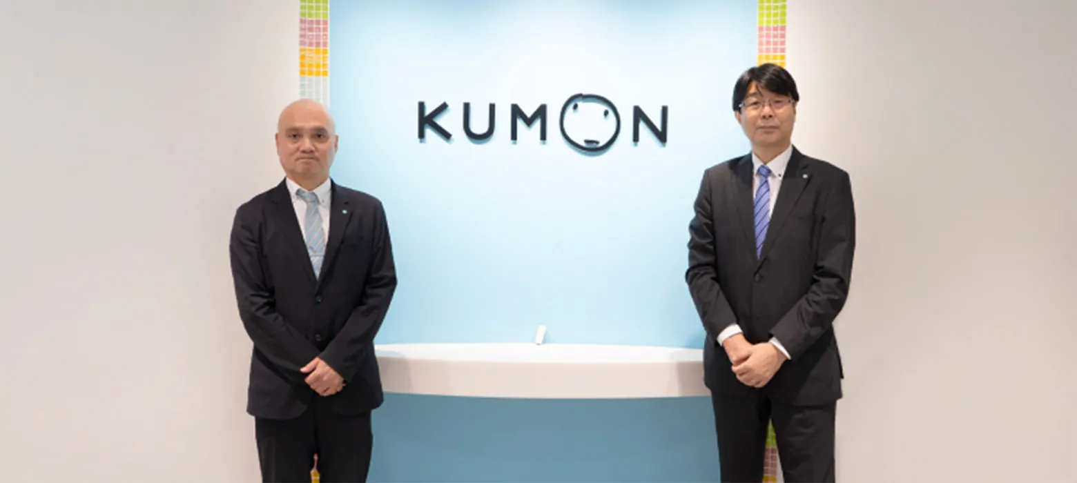 KUMONのロゴを背景に、ダークスーツを着た二名の男性がカウンター両脇でポーズをとっている